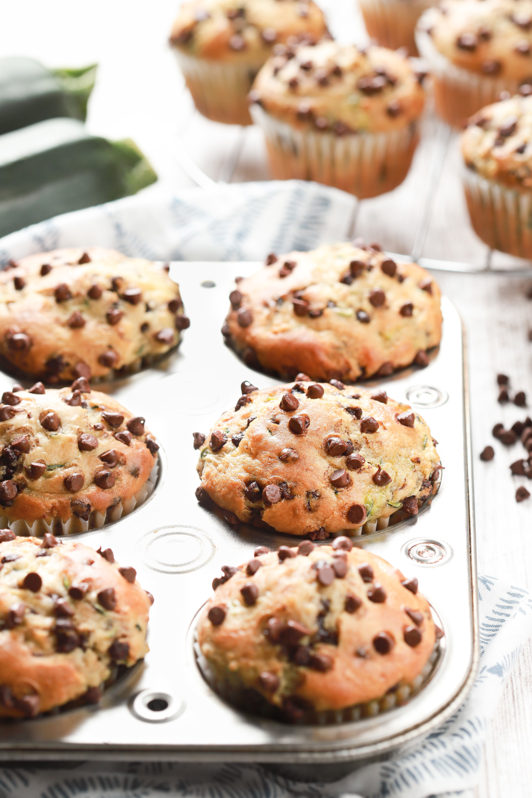 A batch of chocolate chip zucchini muffins in a muffin tin with more muffins on a cooling rack in the background.
