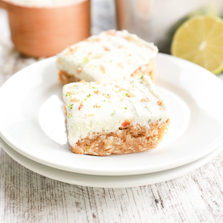 Two lime frosted coconut oat bars on a small white plate with the remaining bars in a baking dish in the background.