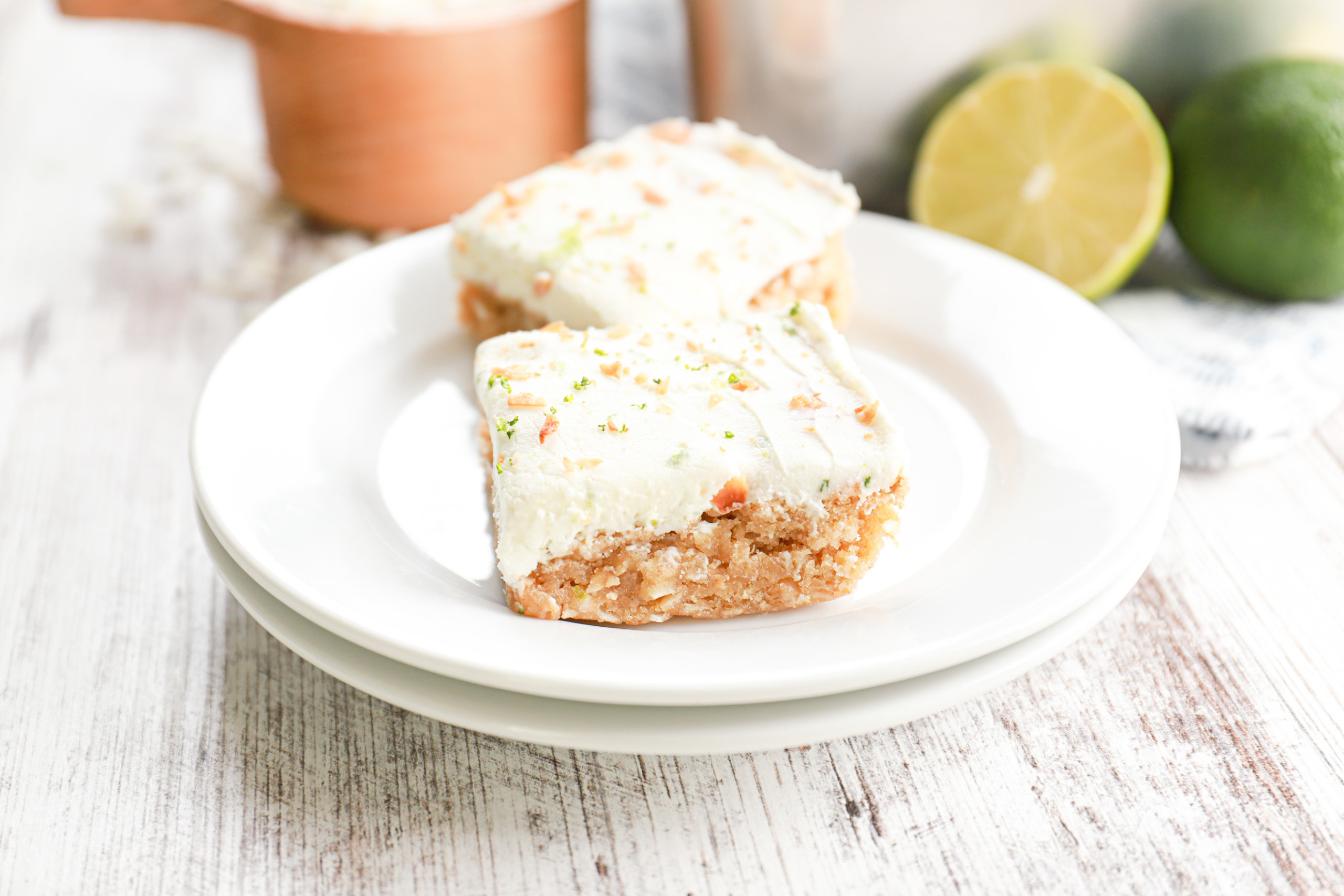 Two lime frosted coconut oat bars on a small white plate with the remaining bars in a baking dish in the background.