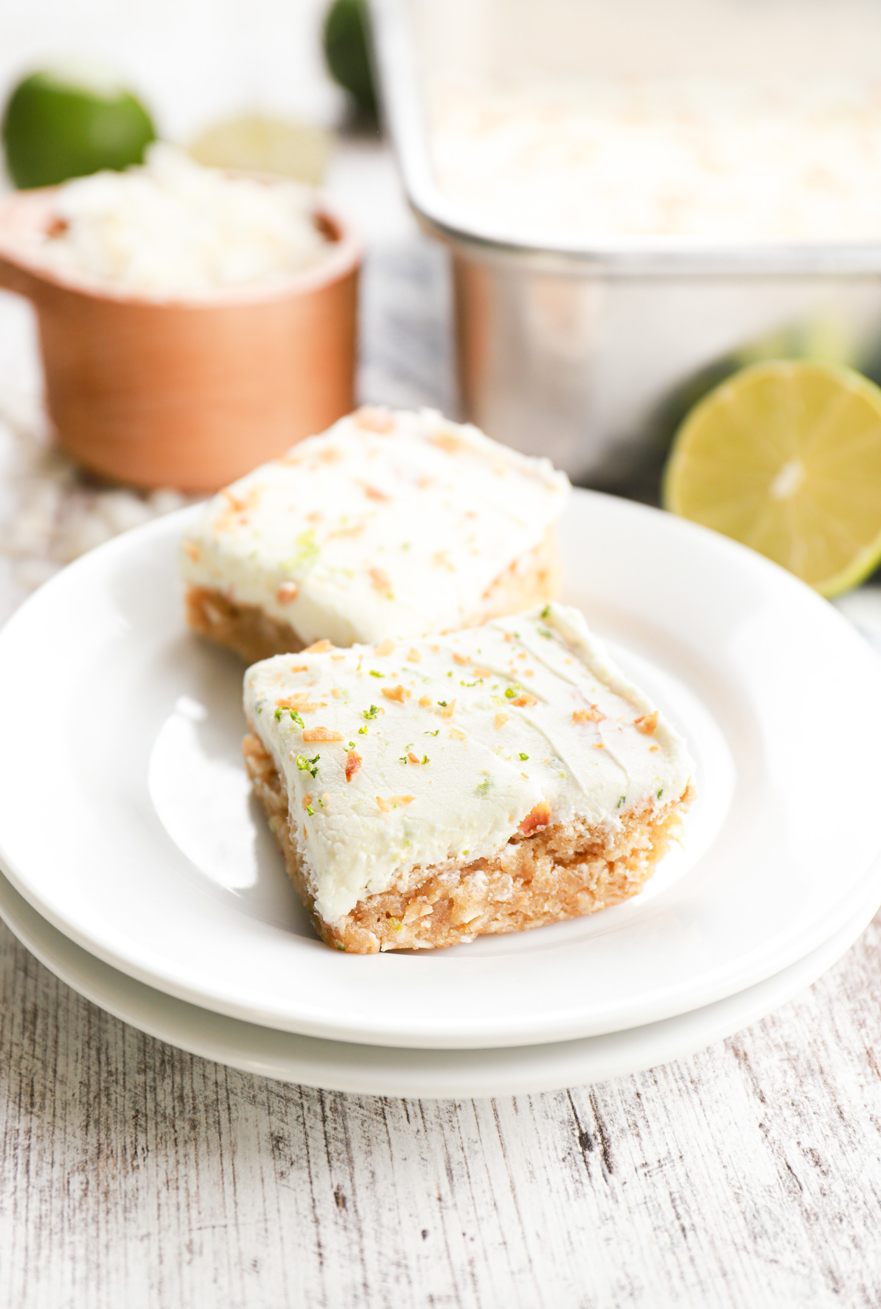 Two lime frosted coconut oat bars on a small white plate with the baking dish with the remainder of the bars in the background.