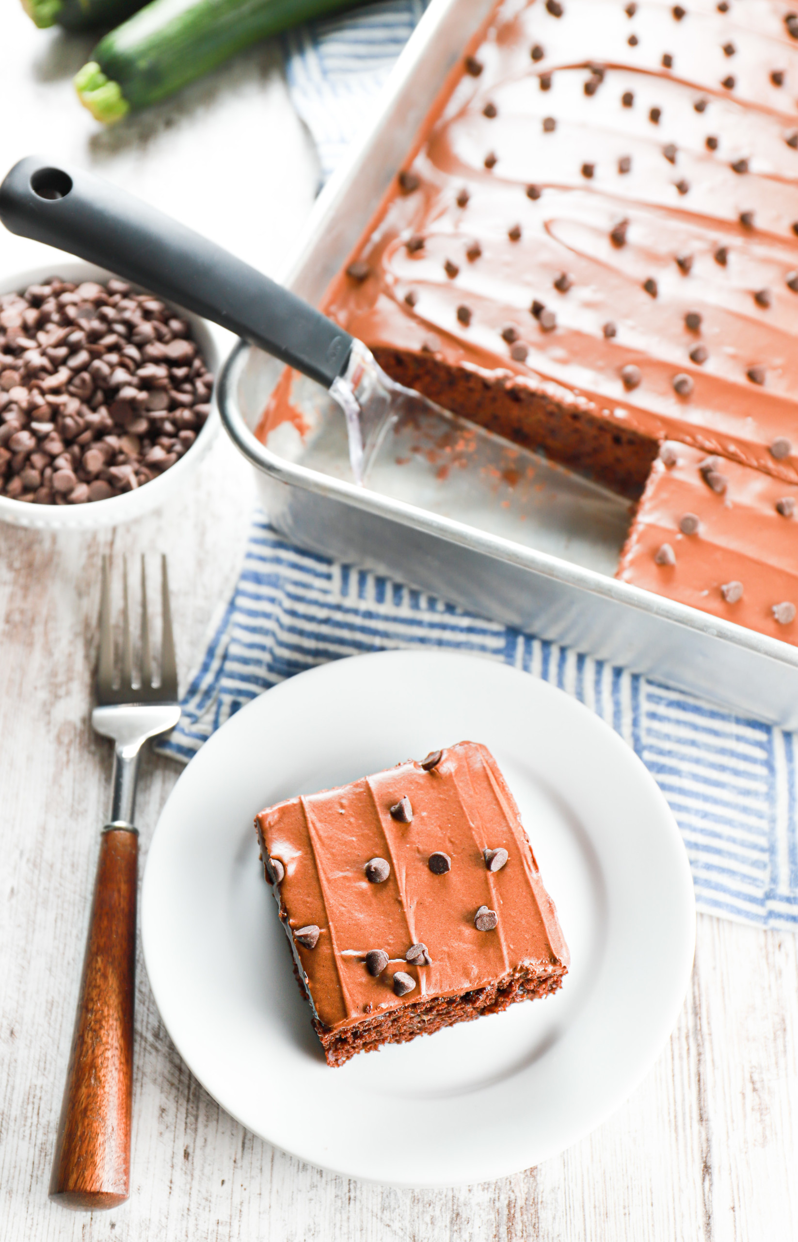 Overhead view of a piece of chocolate frosted zucchini cake on a small white plate and a pan of chocolate frosted zucchini bars.