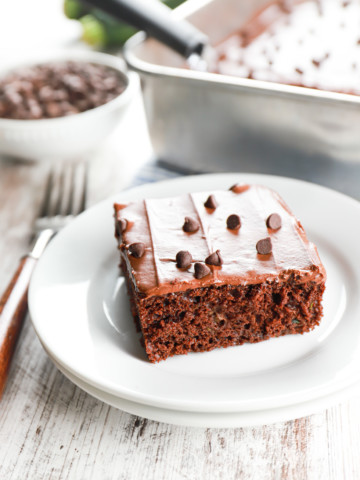 One piece of chocolate frosted zucchini bars on a small white plate with the baking dish full of the remaining bars in the background.