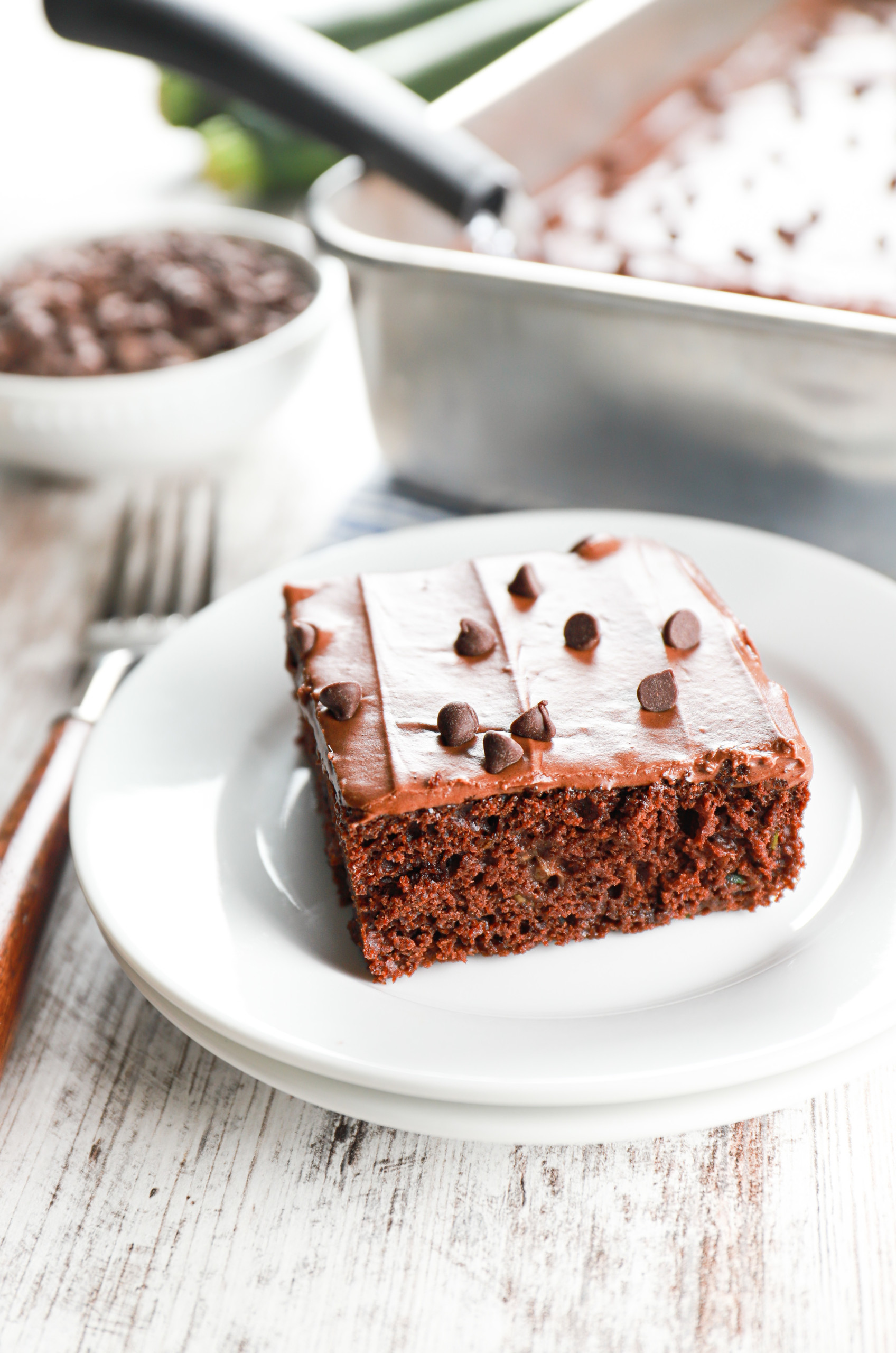 One piece of chocolate frosted zucchini bars on a small white plate with the baking dish full of the remaining bars in the background.