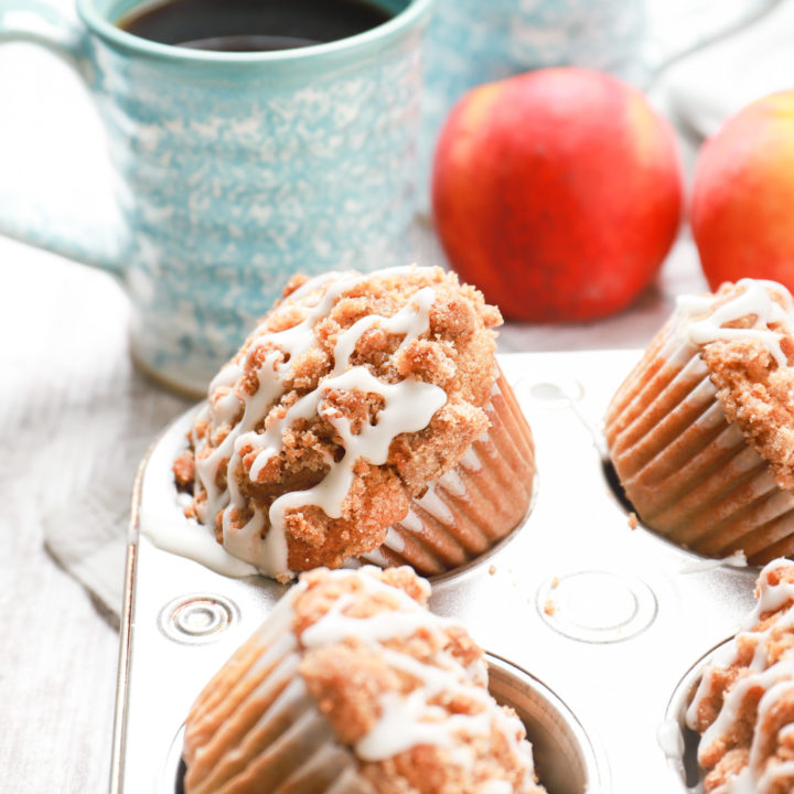Up close side view of an angled peach streusel muffin in a muffin tin with peaches and a blue coffee mug in the background.