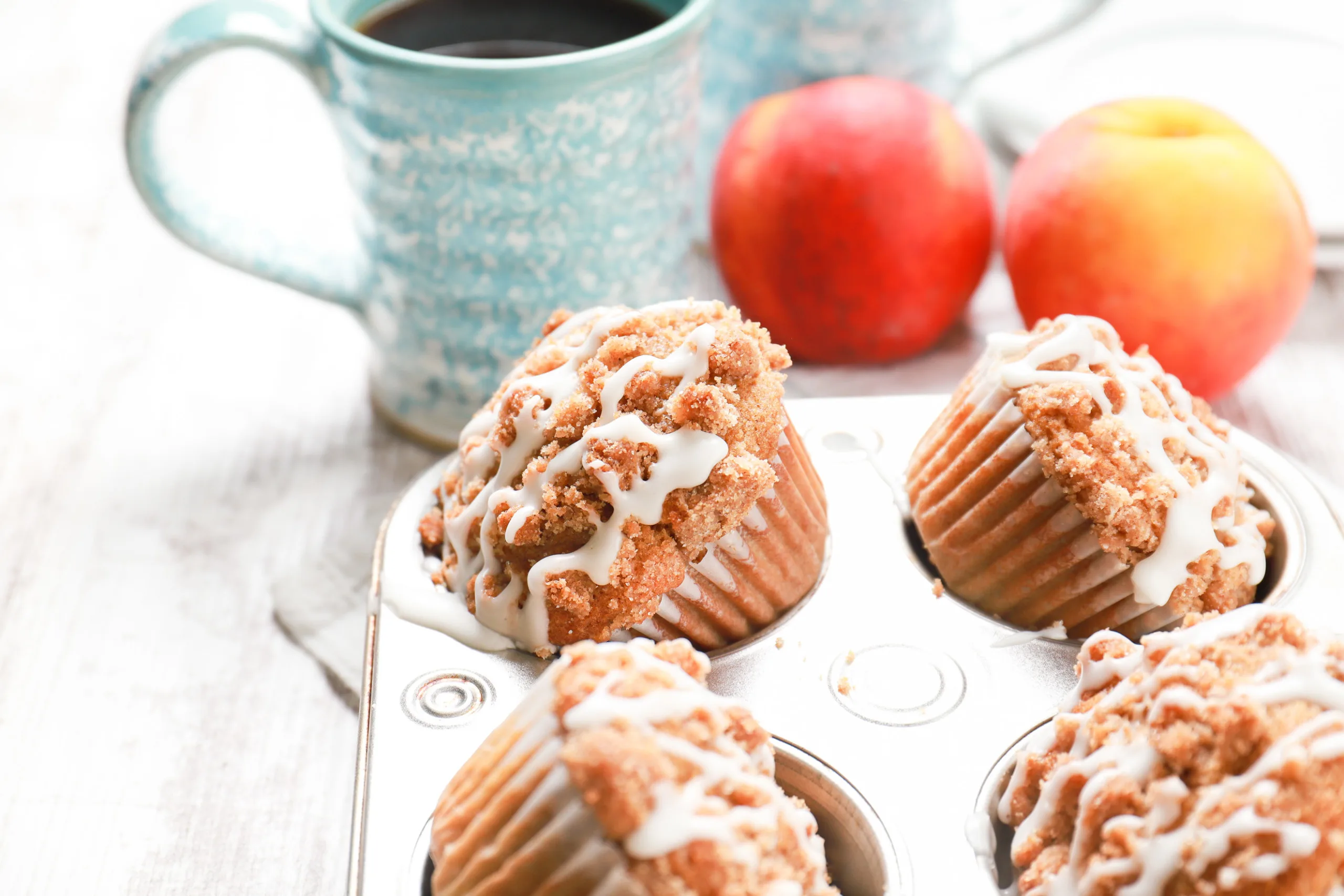 Up close side view of an angled peach streusel muffin in a muffin tin with peaches and a blue coffee mug in the background.