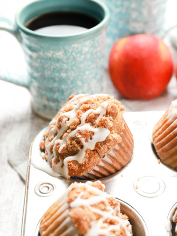 Up close side view of an angled peach coffee cake muffin in a muffin tin with a cup of coffee and couple of peaches in the background.