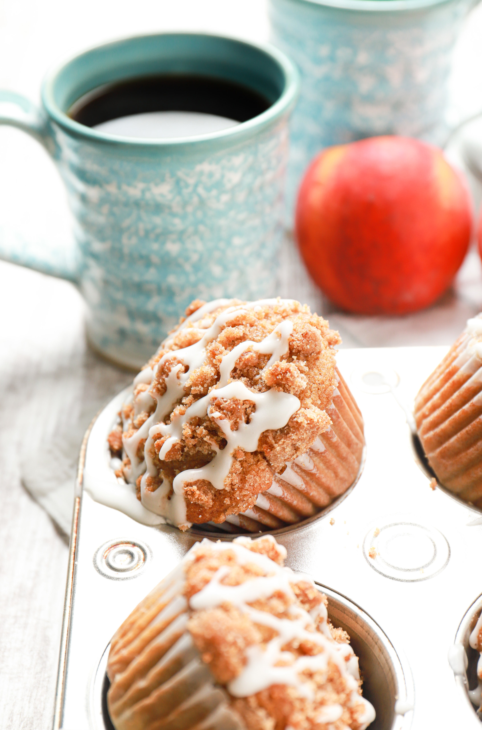 Up close side view of an angled peach coffee cake muffin in a muffin tin with a cup of coffee and couple of peaches in the background.