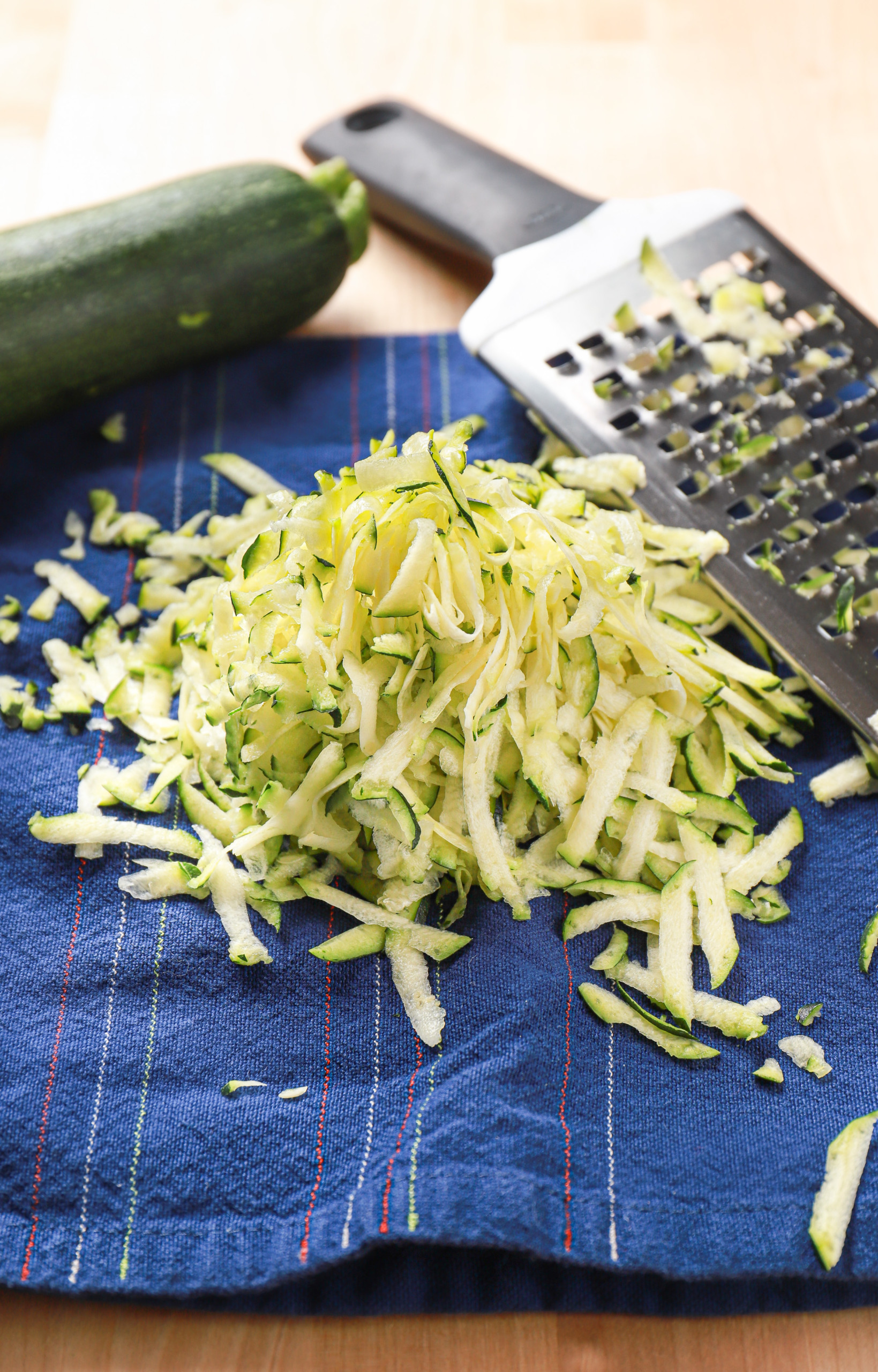 Shredded zucchini on a dark blue towel with a grater and zucchini in the background.