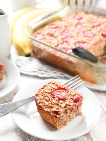 A piece of strawberry banana bread baked oatmeal on a small white plate with a baking dish of the remaining baked oatmeal in the background.