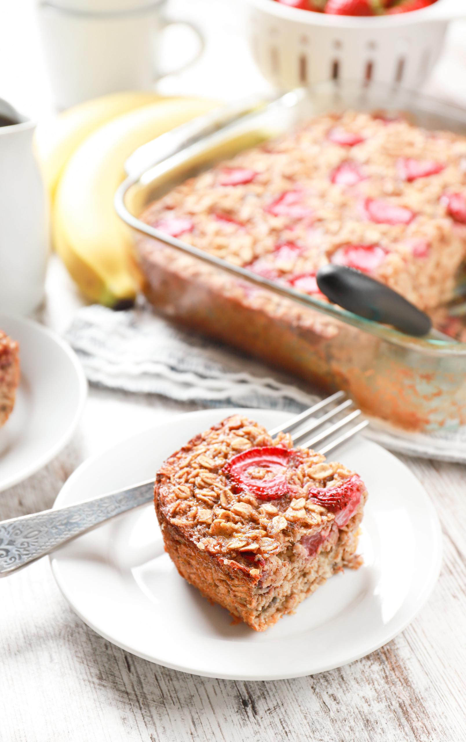 A piece of strawberry banana bread baked oatmeal on a small white plate with a baking dish of the remaining baked oatmeal in the background.