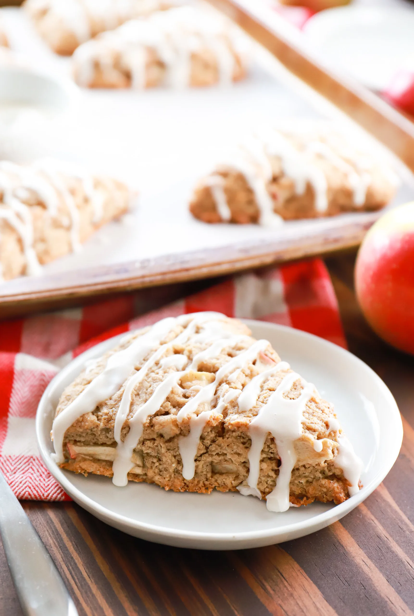 An apple cinnamon scone on a small white plate with remaining scones on a baking sheet in the background.