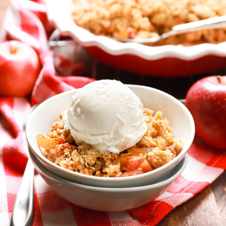 Side view of a white bowl full of caramel apple crisp topped with vanilla ice cream with the remainder of the apple crisp in the background.