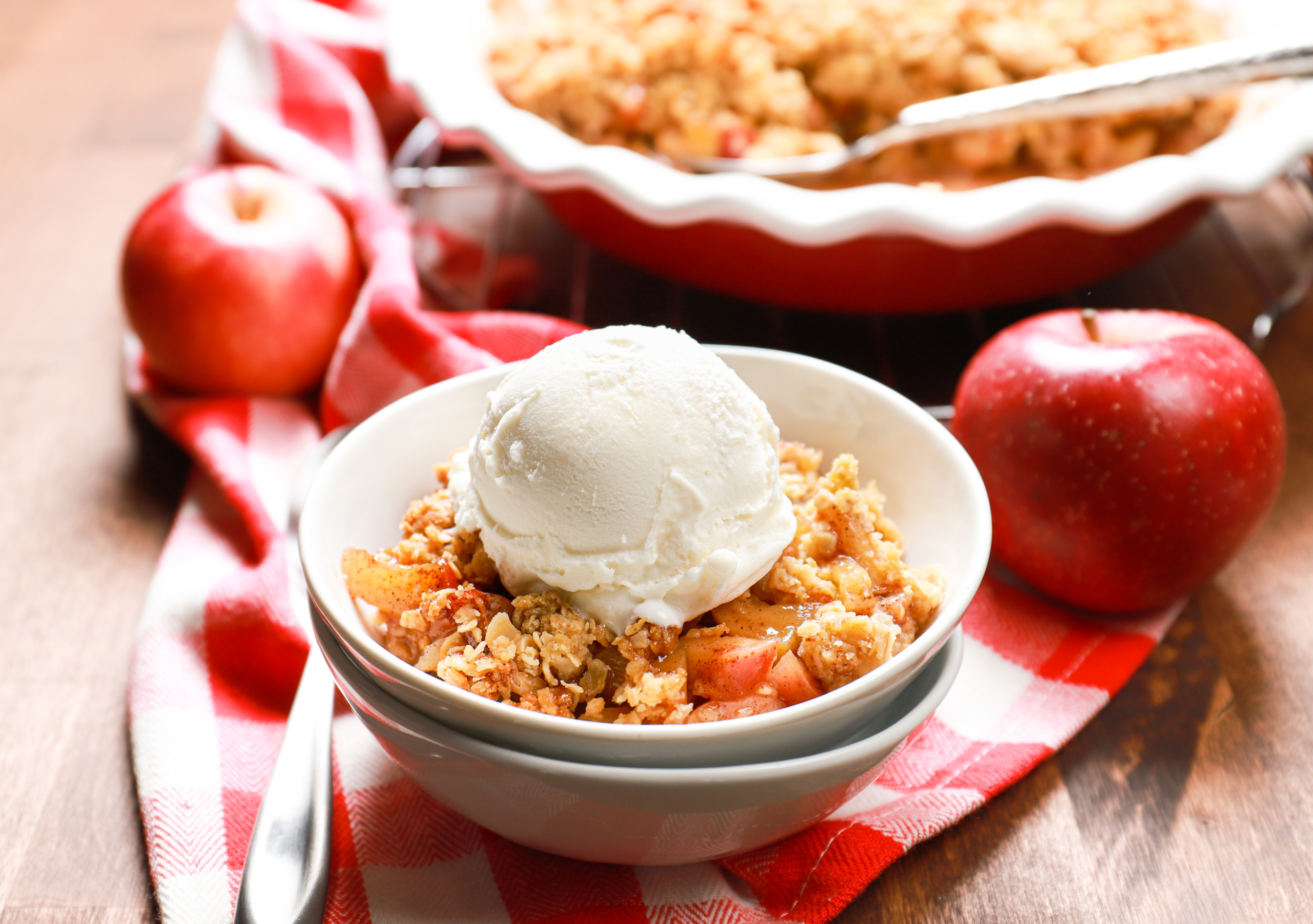 Side view of a white bowl full of caramel apple crisp topped with vanilla ice cream with the remainder of the apple crisp in the background.