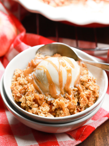 A small white bowl filled with caramel apple crisp topped with vanilla ice cream and a drizzle of caramel sauce.