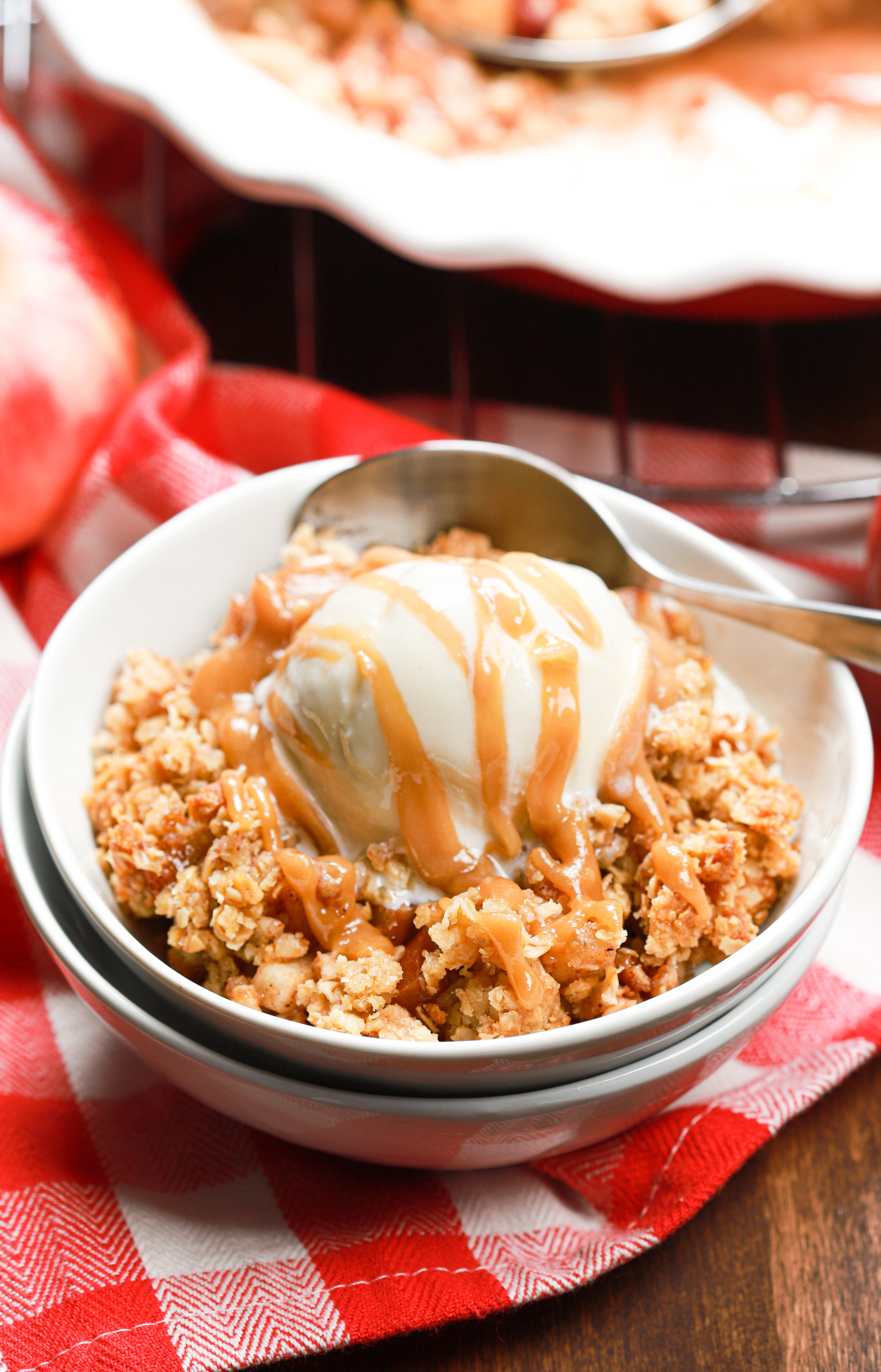 A small white bowl filled with caramel apple crisp topped with vanilla ice cream and a drizzle of caramel sauce.