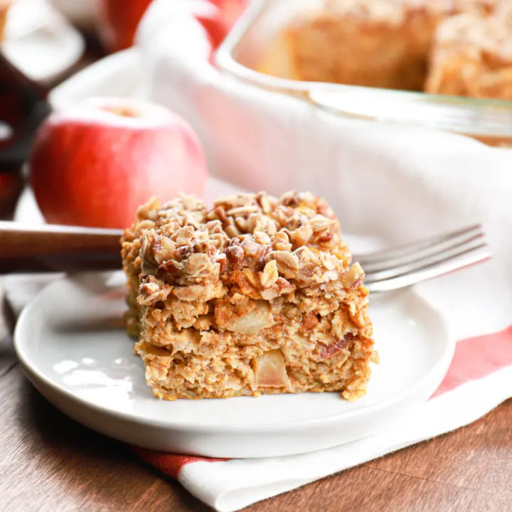 A piece of pumpkin apple baked oatmeal on a small white plate with the remaining oatmeal in a baking dish in the background.