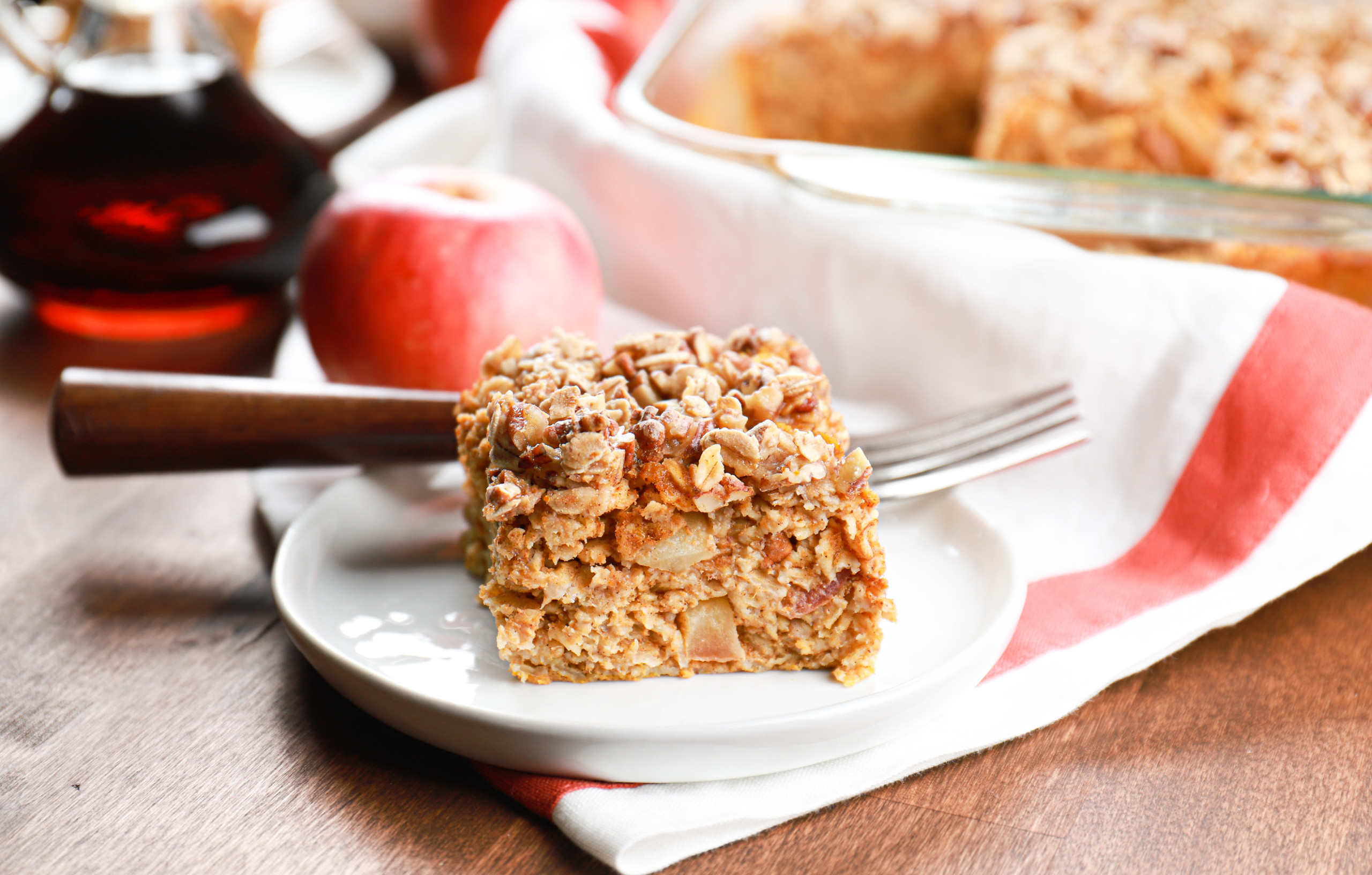 A piece of pumpkin apple baked oatmeal on a small white plate with the remaining oatmeal in a baking dish in the background.