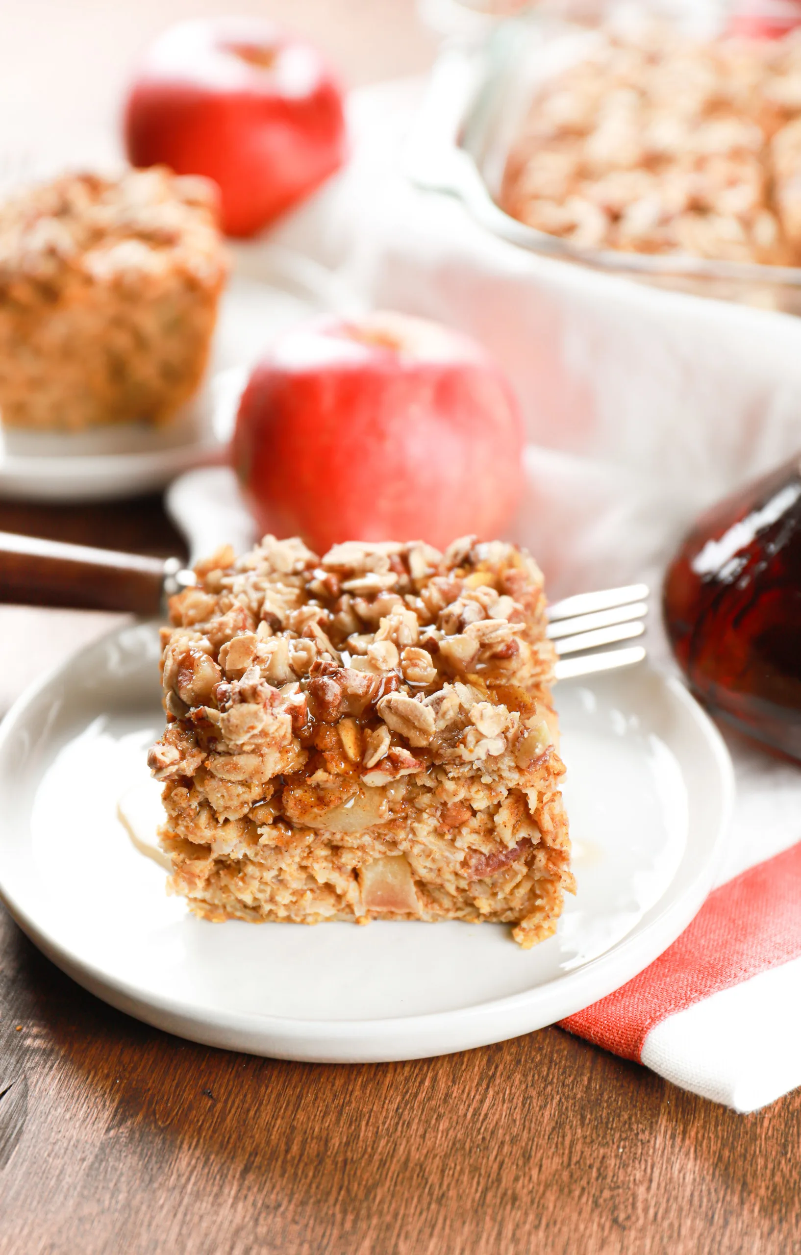 A slice of pumpkin apple baked oatmeal that has been covered in maple syrup on a small white plate with the remaining oatmeal in the background.