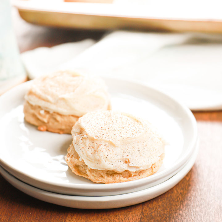 Up close view of two frosted eggnog cookies on a small white plate with more cookies on a baking sheet in the background.