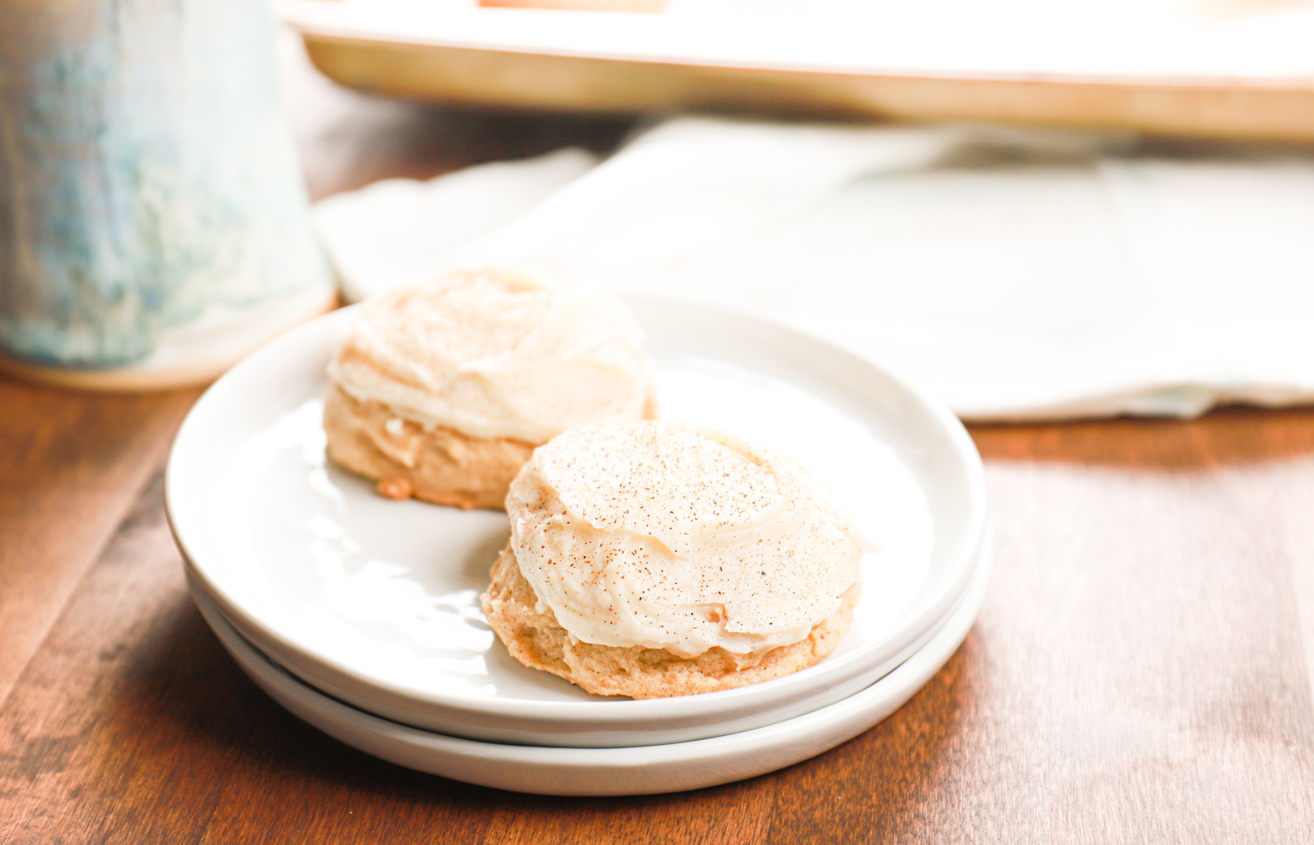 Up close view of two frosted eggnog cookies on a small white plate with more cookies on a baking sheet in the background.