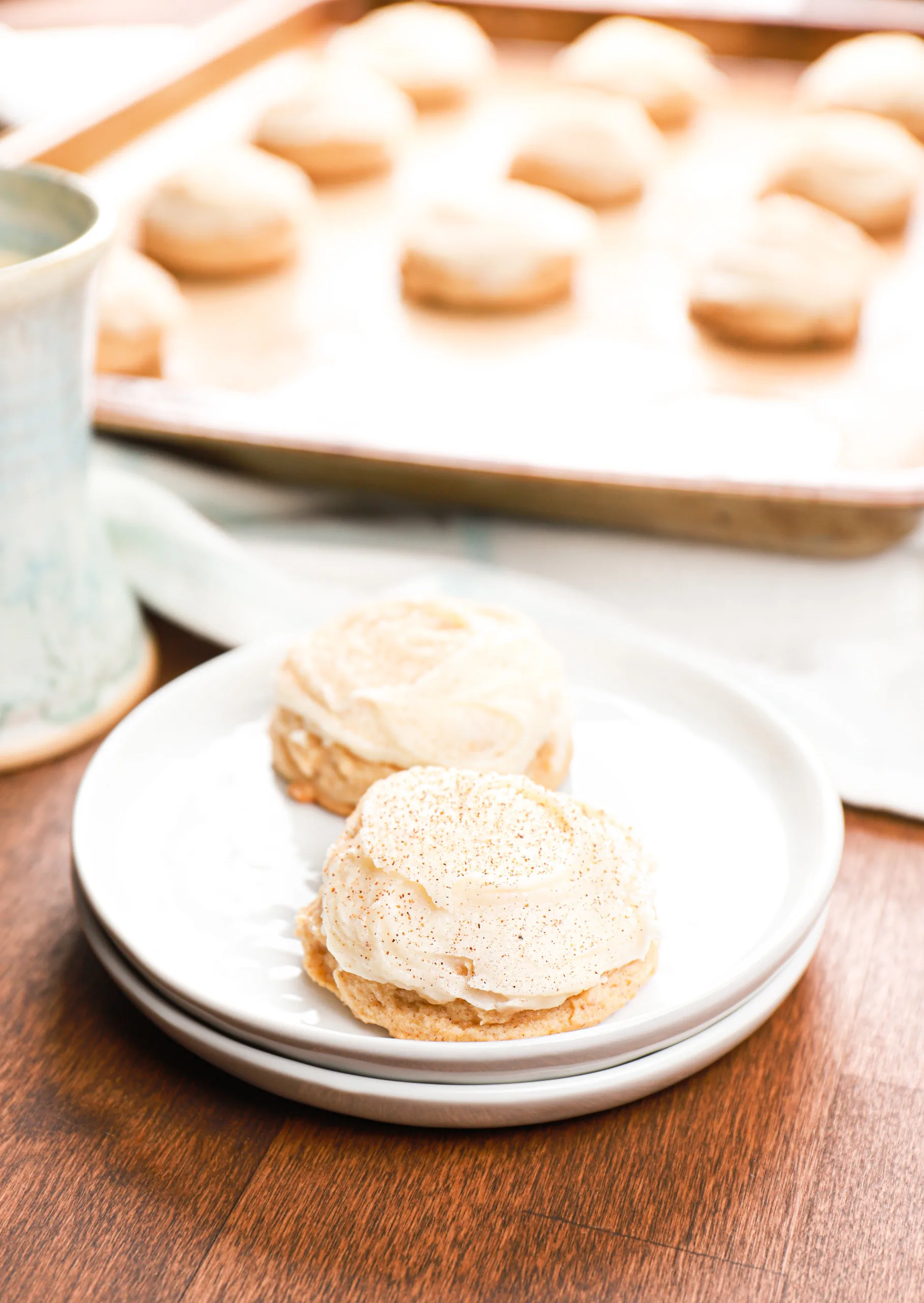 Two frosted eggnog cookies on a small white plate with the remainder of the batch on a baking sheet in the background.