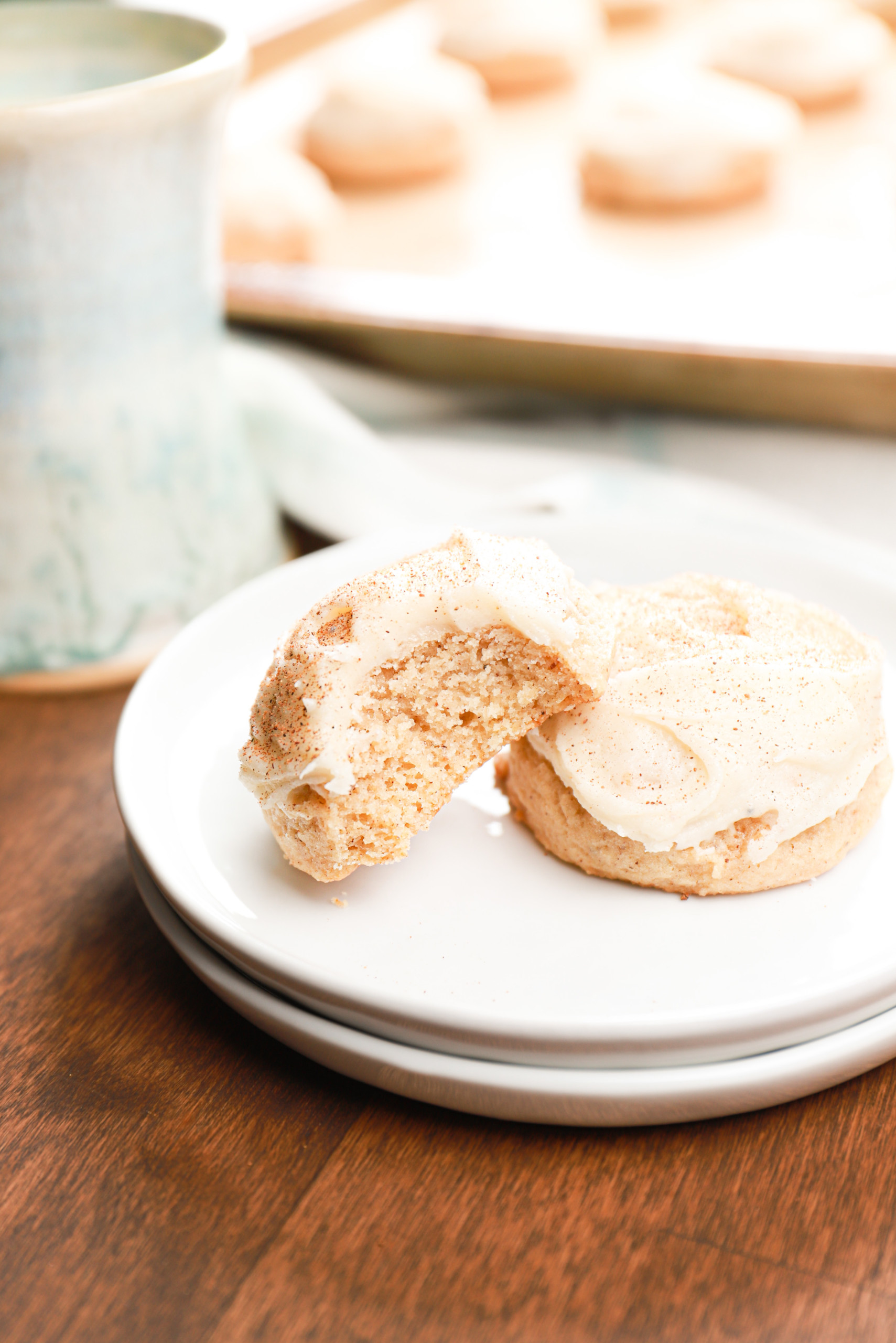 Two frosted eggnog cookies with one having a bite taken out of it on a small white plate with the remaining cookies on a baking sheet in the background.