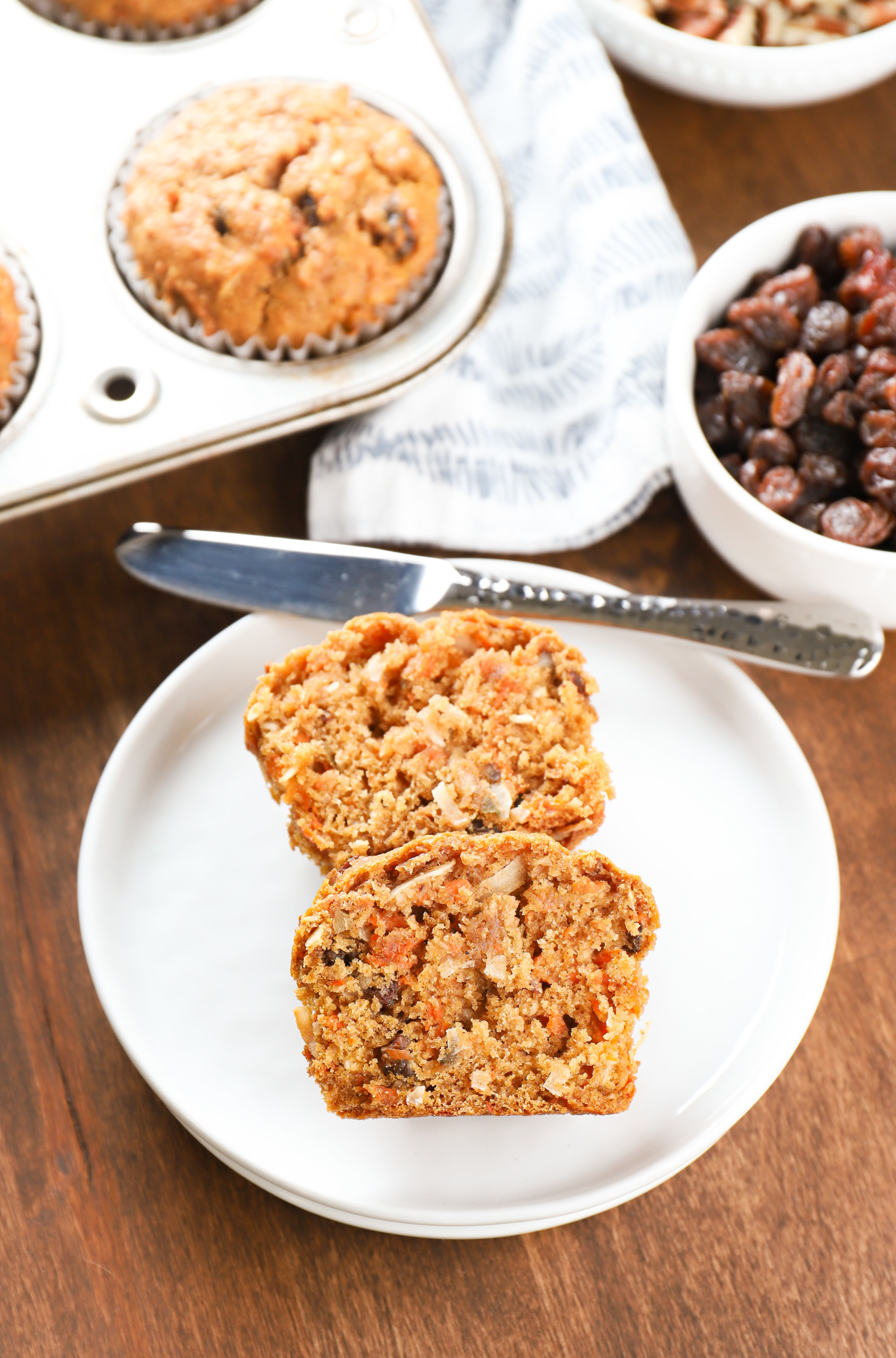 Overhead view of a morning glory muffin sliced in half on a small white plate with a muffin tin with the remaining muffins in the background.