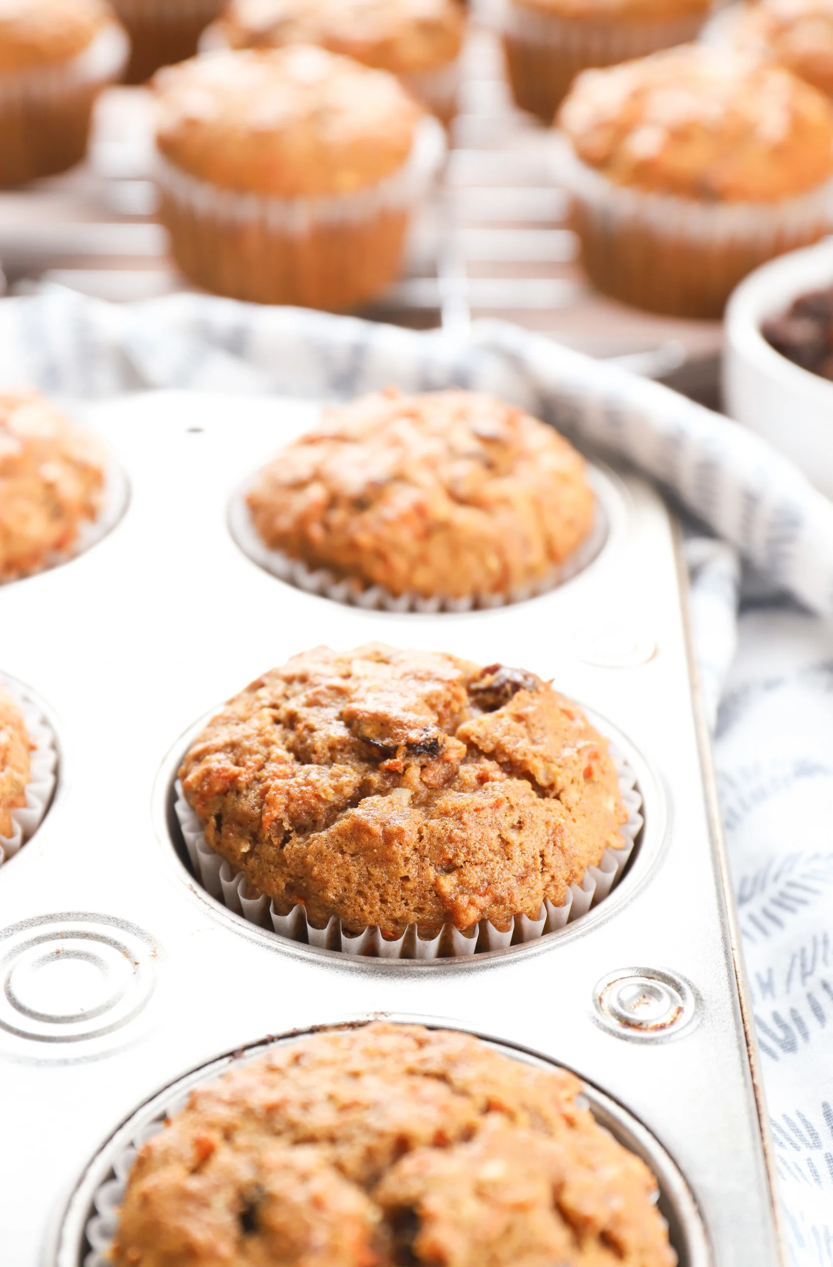 Side view of the top of a morning glory muffin in an aluminum muffin tin with more muffins on a wire cooling rack in the background.