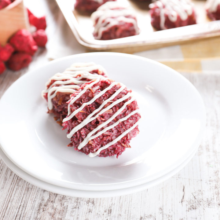 Two coconut raspberry macaroons on a small white plate with the remaining macaroons in the background on a baking sheet.