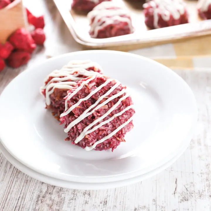 Two coconut raspberry macaroons on a small white plate with the remaining macaroons in the background on a baking sheet.