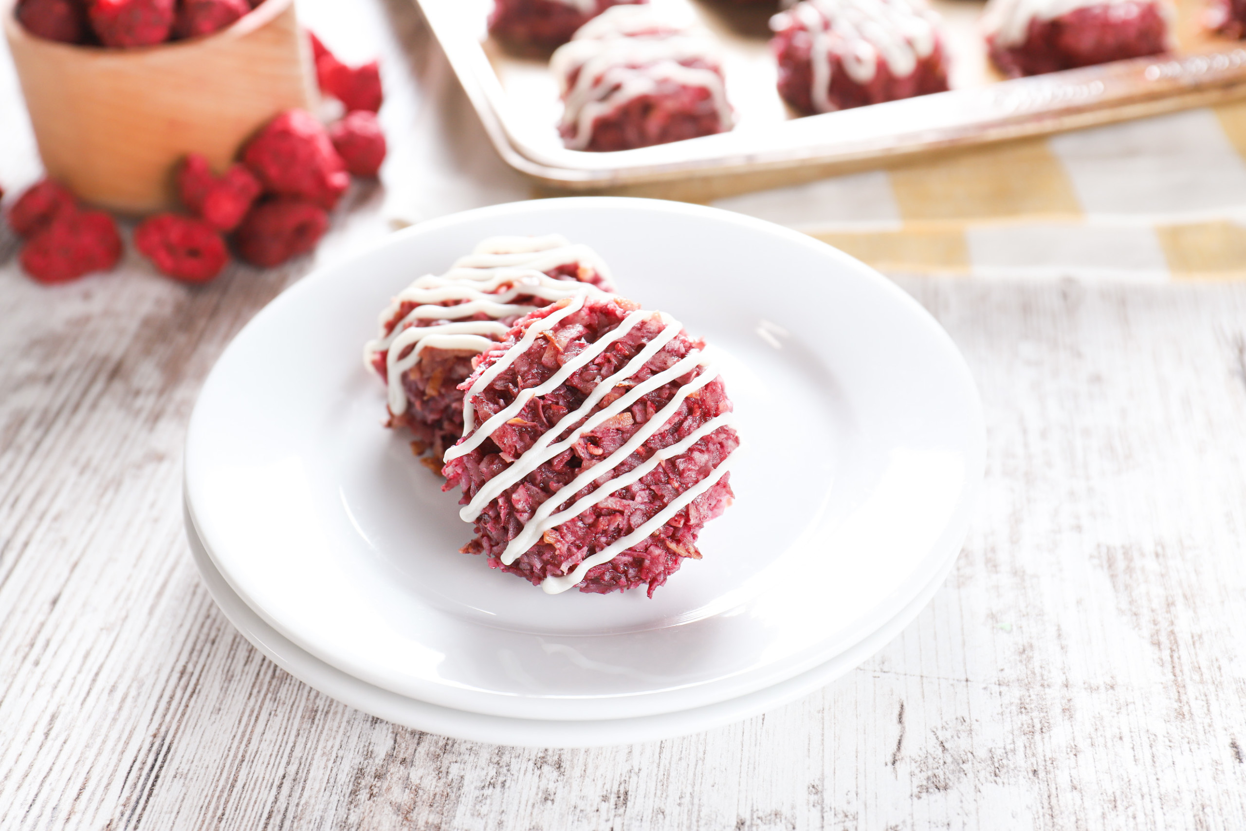 Two coconut raspberry macaroons on a small white plate with the remaining macaroons in the background on a baking sheet.