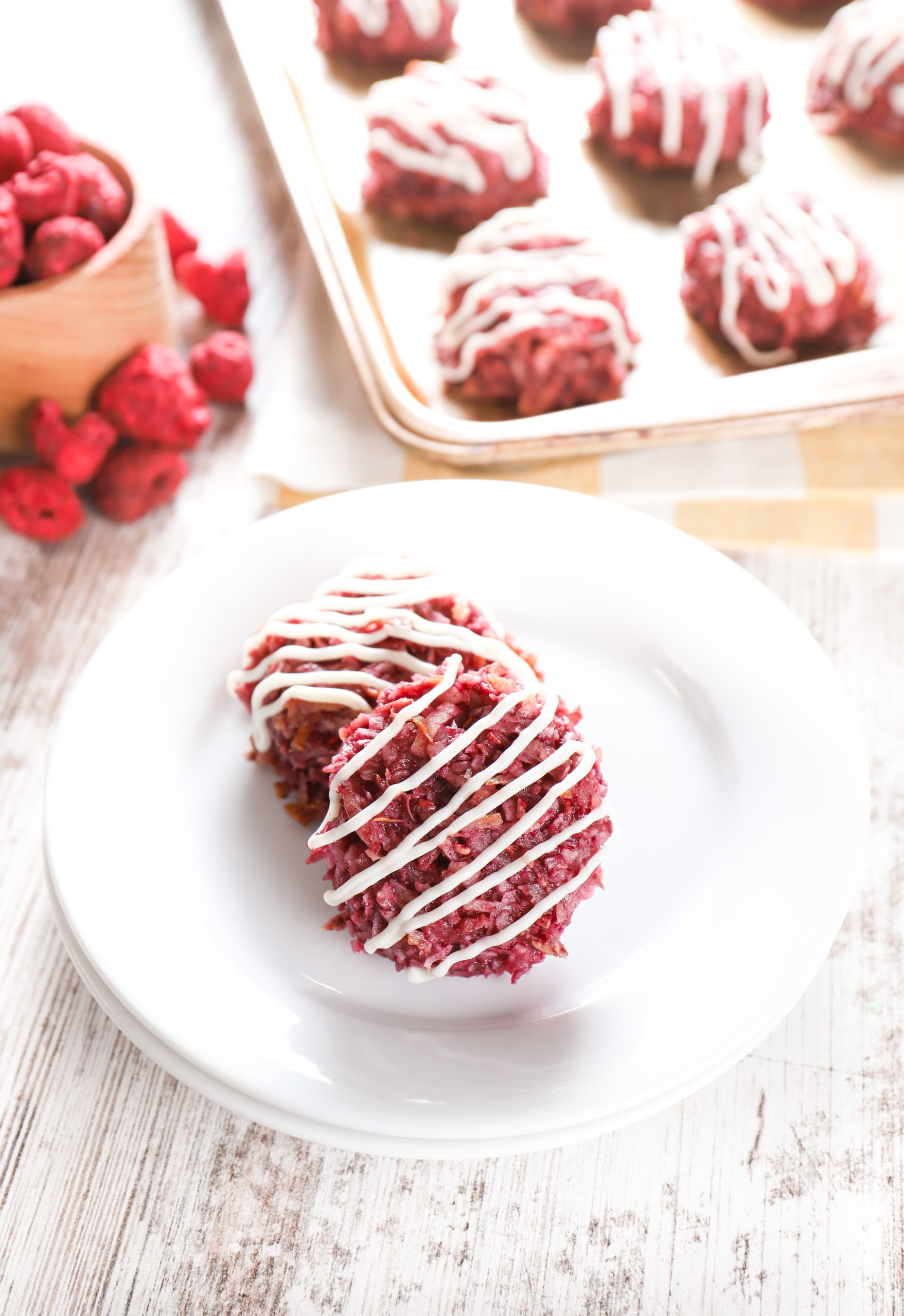 Overhead view of two raspberry coconut macaroons on a small white plate with the remainder of the batch on a baking sheet in the background.