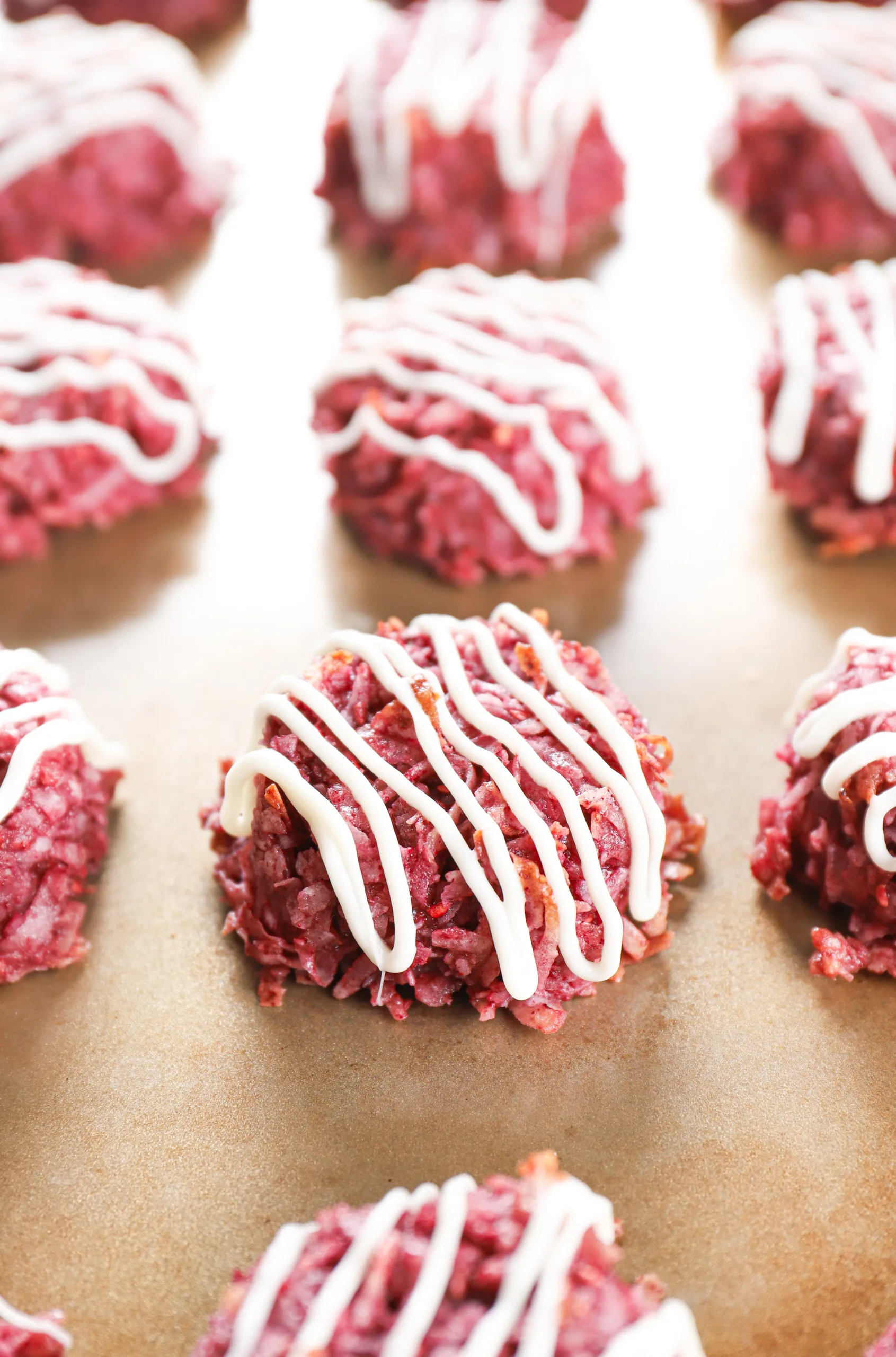 Side view of a white chocolate drizzled raspberry coconut macaroon on an aluminum baking sheet.