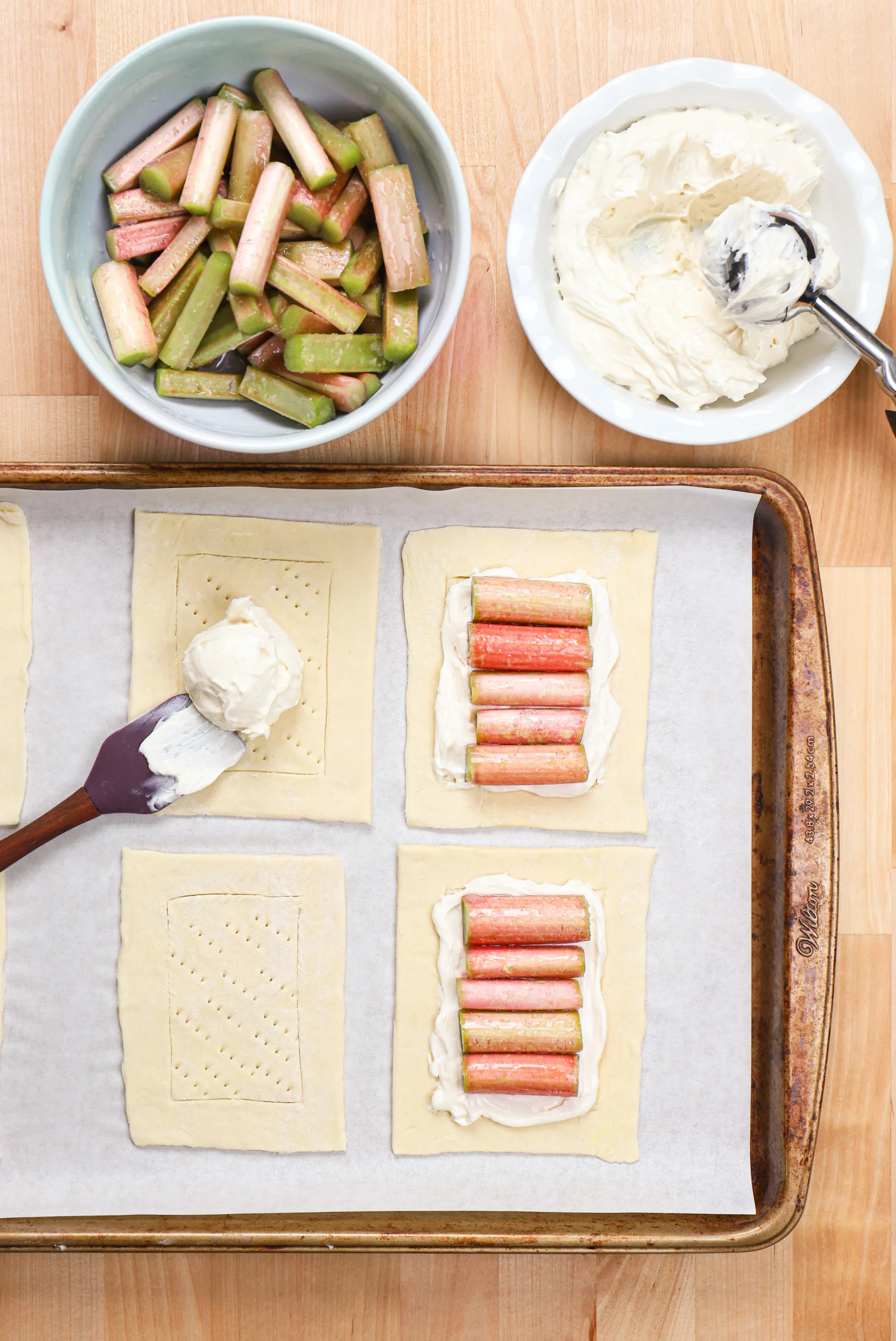 Pieces of puff pastry getting topped with cream cheese and pieces of rhubarb.