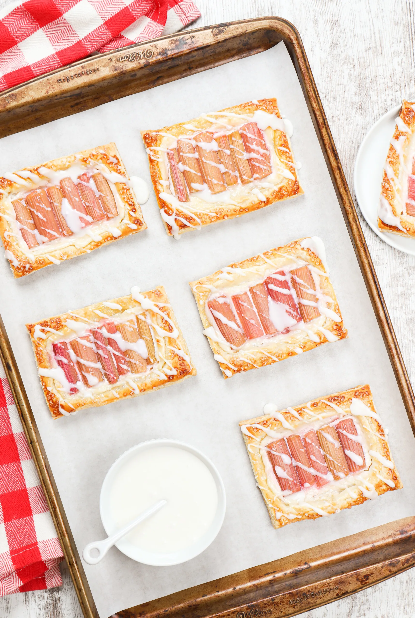 Overhead view of a batch of rhubarb cream cheese danishes on a baking sheet getting drizzled with a confectioners sugar glaze.