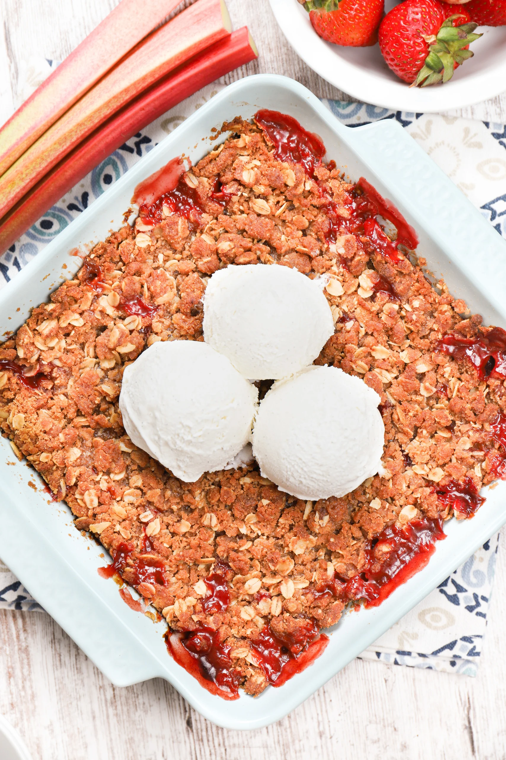 Overhead view of a batch of strawberry rhubarb crisp in a light blue dish with three scoops of vanilla ice cream on top.