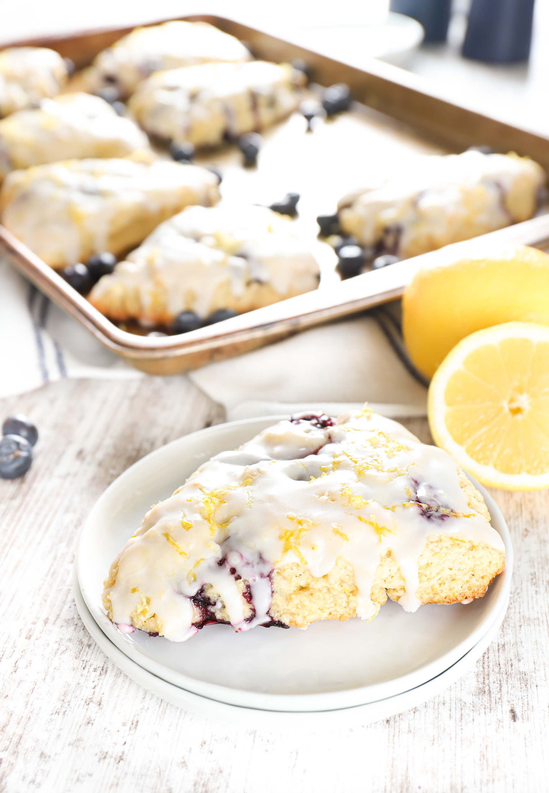 A lemon blueberry scone on a small white plate with a pan of scones in the background.
