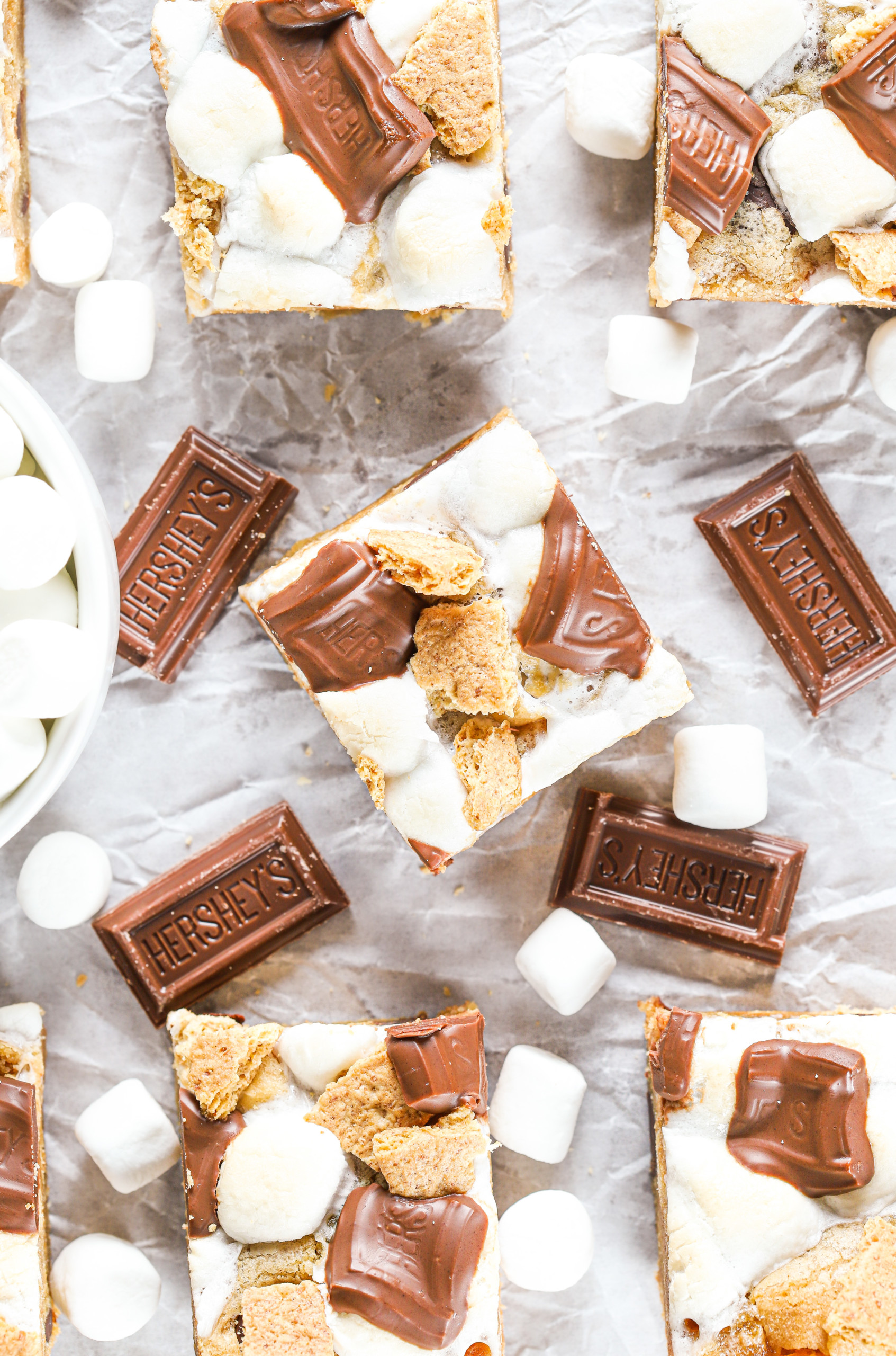 Up close overhead view of a peanut butter smores cookie bar on a piece of parchment paper surrounded by chocolate pieces, mini marshmallows, and the remaining bars.