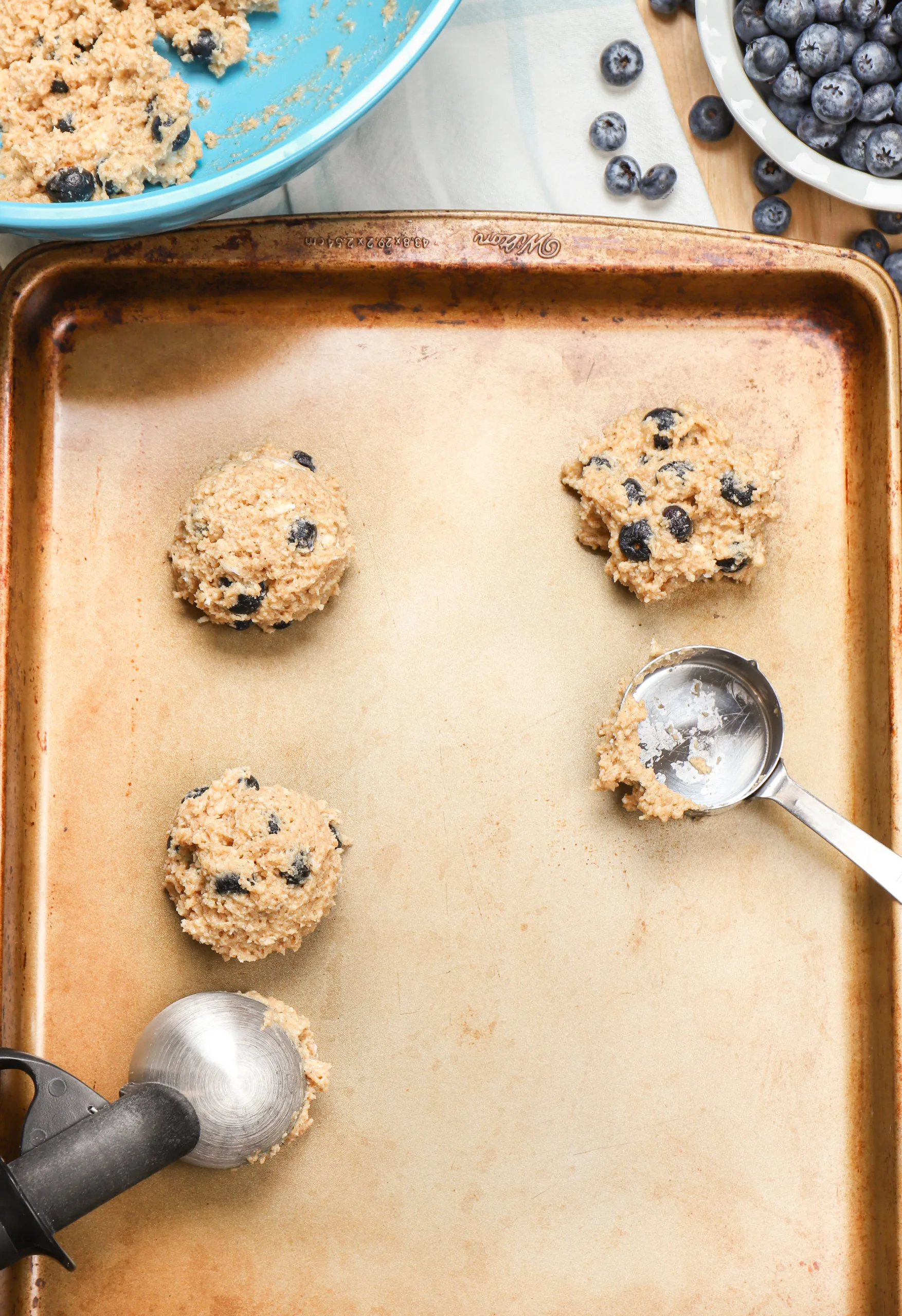 Overhead view of putting blueberry fritter dough onto an aluminum baking sheet using an ice cream scoop and a measuring cup.