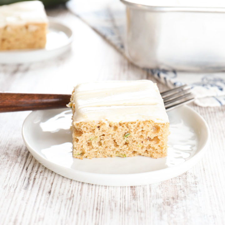 Side view of a zucchini bar on a small white plate with another plate with a zucchini bar and the remaining pan of bars in the background.