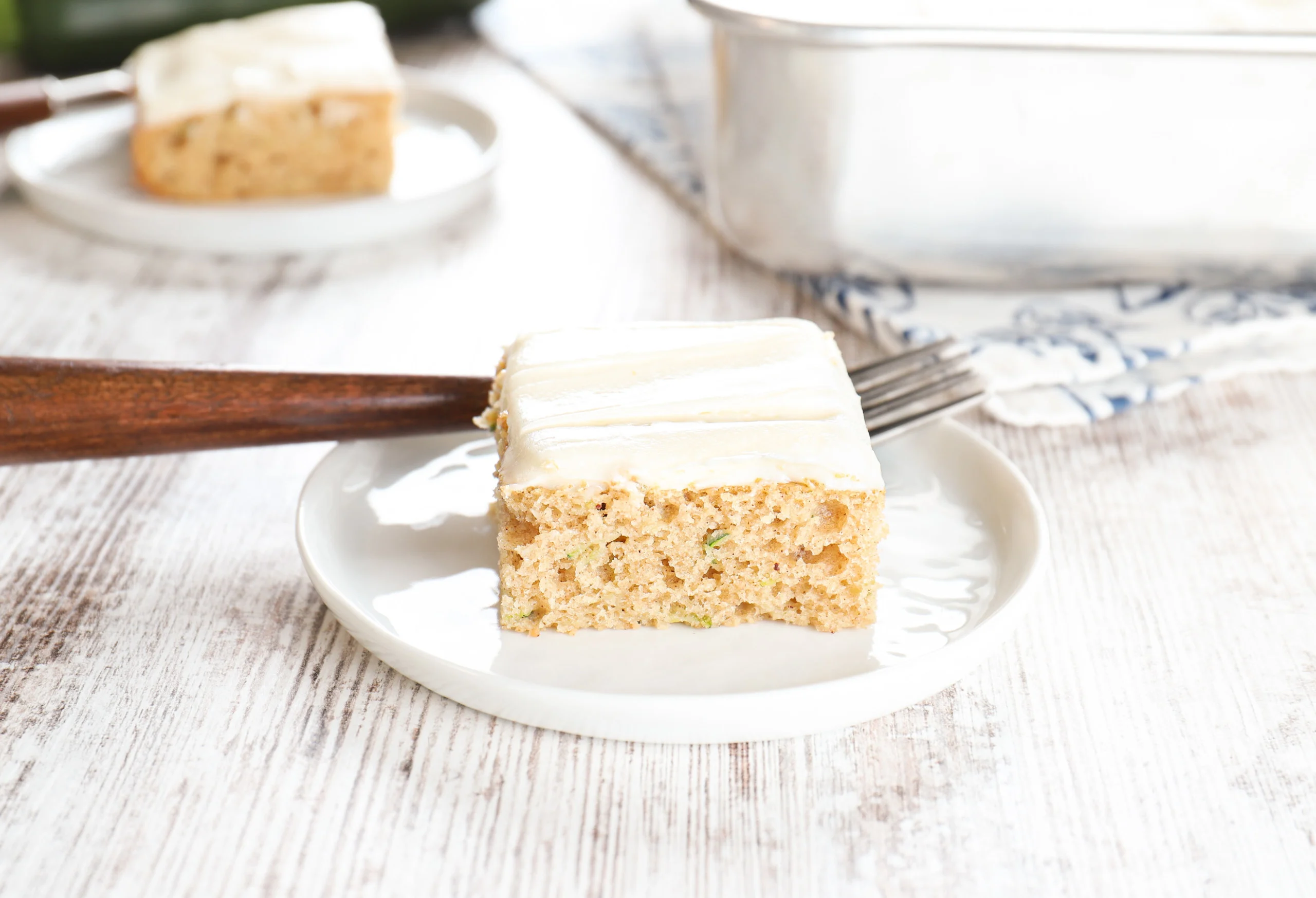 Side view of a zucchini bar on a small white plate with another plate with a zucchini bar and the remaining pan of bars in the background.