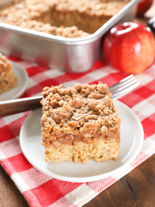 One piece of apple streusel baked oatmeal on a small white plate with the remaining baking dish of oatmeal in the background.