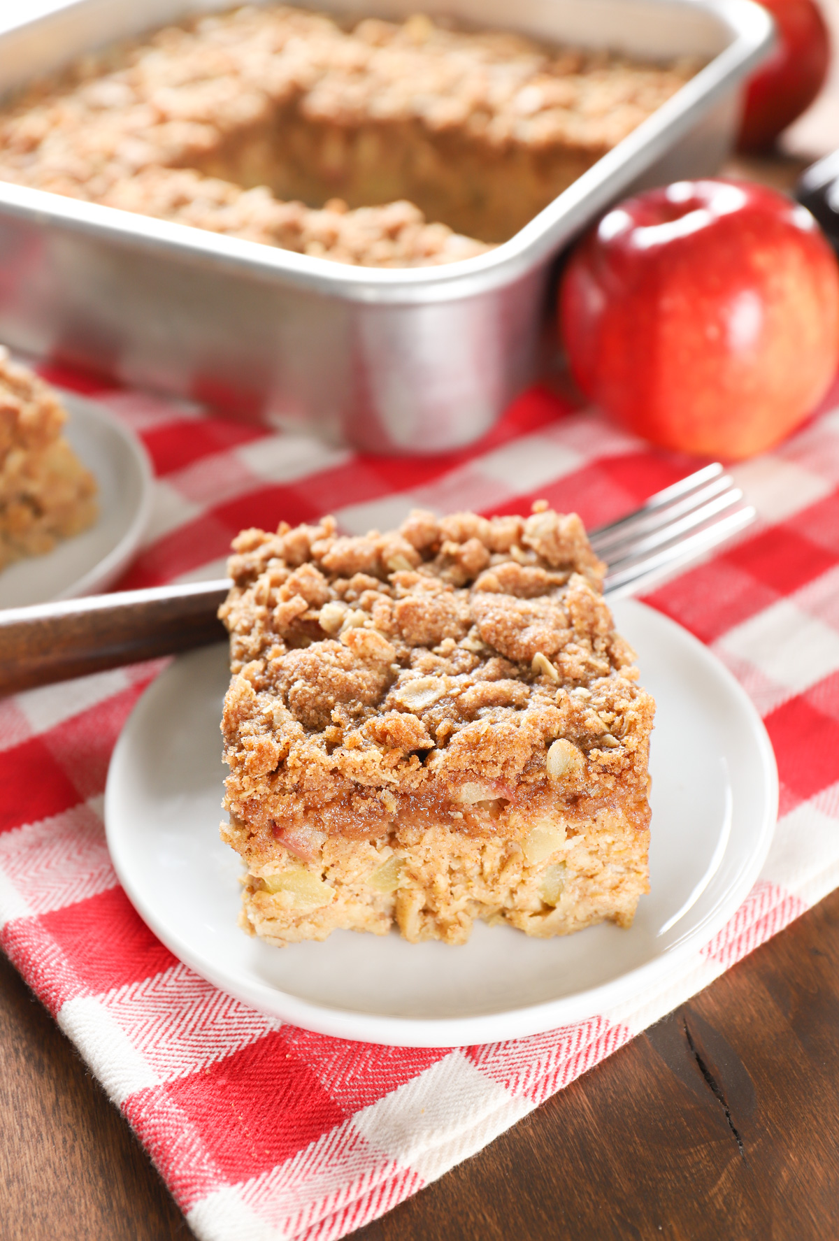 One piece of apple streusel baked oatmeal on a small white plate with the remaining baking dish of oatmeal in the background.